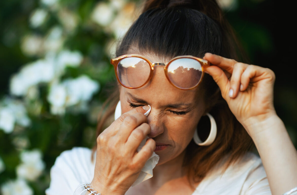 Summer time. sad woman in white shirt with handkerchief and eyeglasses dealing with summer eye allergies near a flowering tree.