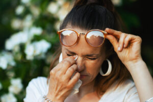 Summer time. sad woman in white shirt with handkerchief and eyeglasses dealing with summer eye allergies near a flowering tree.