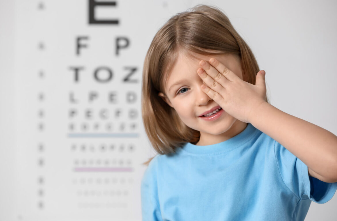 Little girl getting a pediatric eye exam; covering her eye against vision test chart