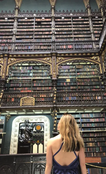 Our-Story-1-370x607 A woman in a blue, backless dress stands in a grand, ornate library with tall, gilded bookshelves and intricate metalwork, facing a statue near a decorative doorway.